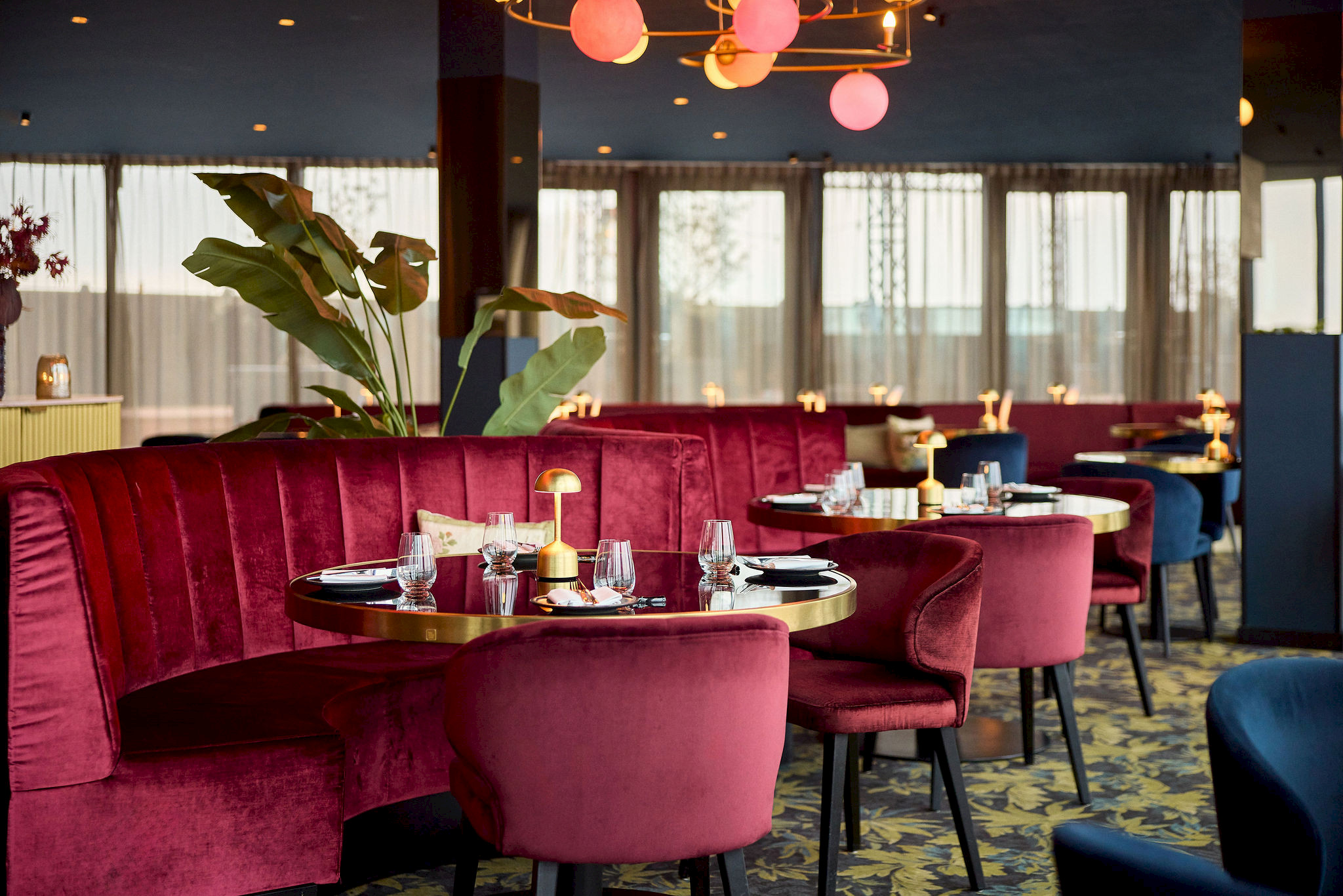Brede shot van het Mr. Sato restaurantinterieur met velvet fauteuils, gouden tafels en gordijnen voor grote ramen
