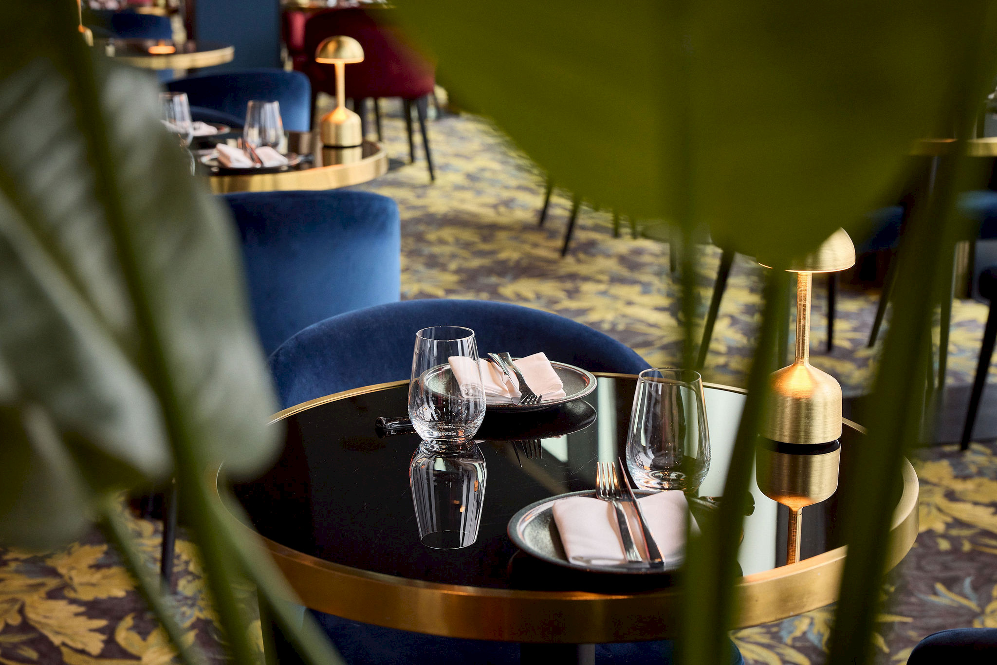 Close-up van een gedekte tafel met gouden rand, waterglazen en roze servetten, omlijst door tropische bladeren