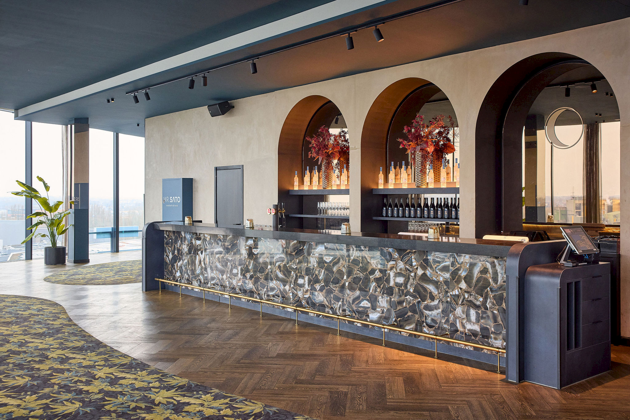 Lange skybar met onyx-marmeren counter en gouden lijst, hoge bogen en daglicht door panoramaramen