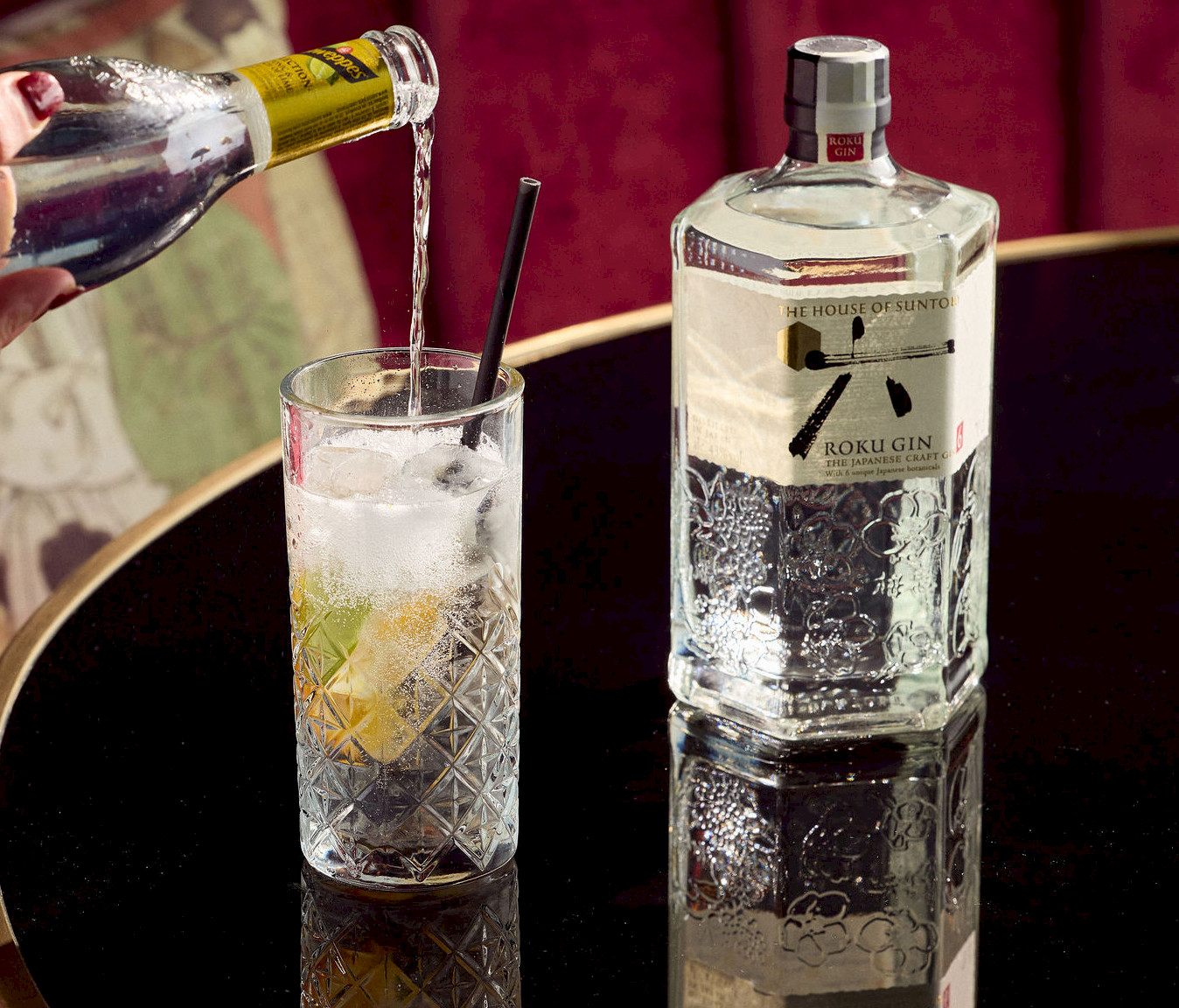 Kristallen tumbler met ijs en limoen waarin Roku Japanese craft gin wordt geschonken naast de sierlijke fles op een gouden tafel